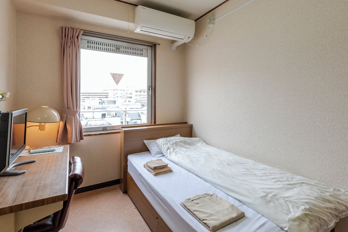 Habitación de hotel compacta con una cama individual, un escritorio con lámpara, televisión y ventana con cortinas.