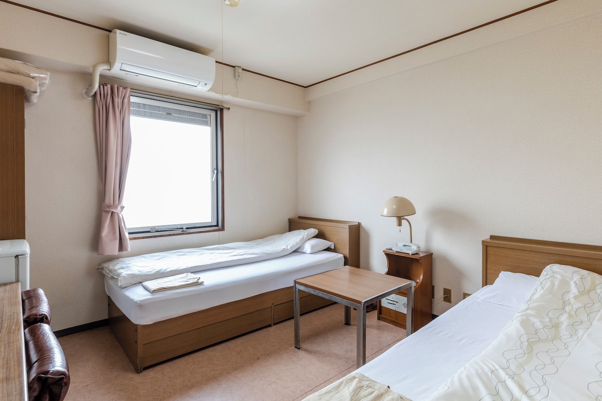 A hotel room with a single bed, a bedside table, a lamp, a window with curtains, and an air conditioning unit.
