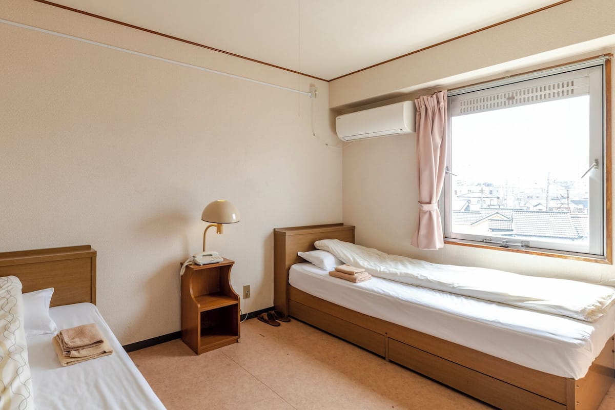 Habitación de hotel con dos camas, una mesita de noche con lámpara, un equipo de aire acondicionado y una ventana con vistas a edificios.