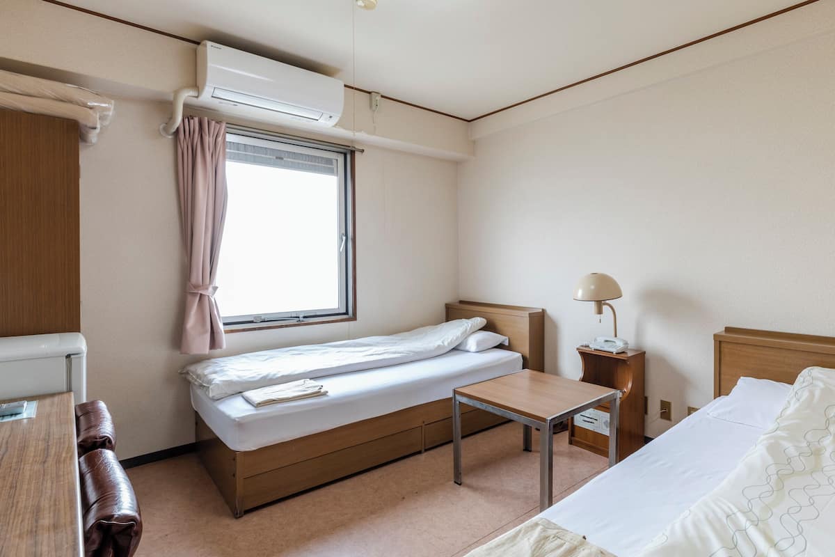 Habitación de hotel con una cama individual, una mesita de noche, una lámpara, una ventana con cortinas y un equipo de aire acondicionado.