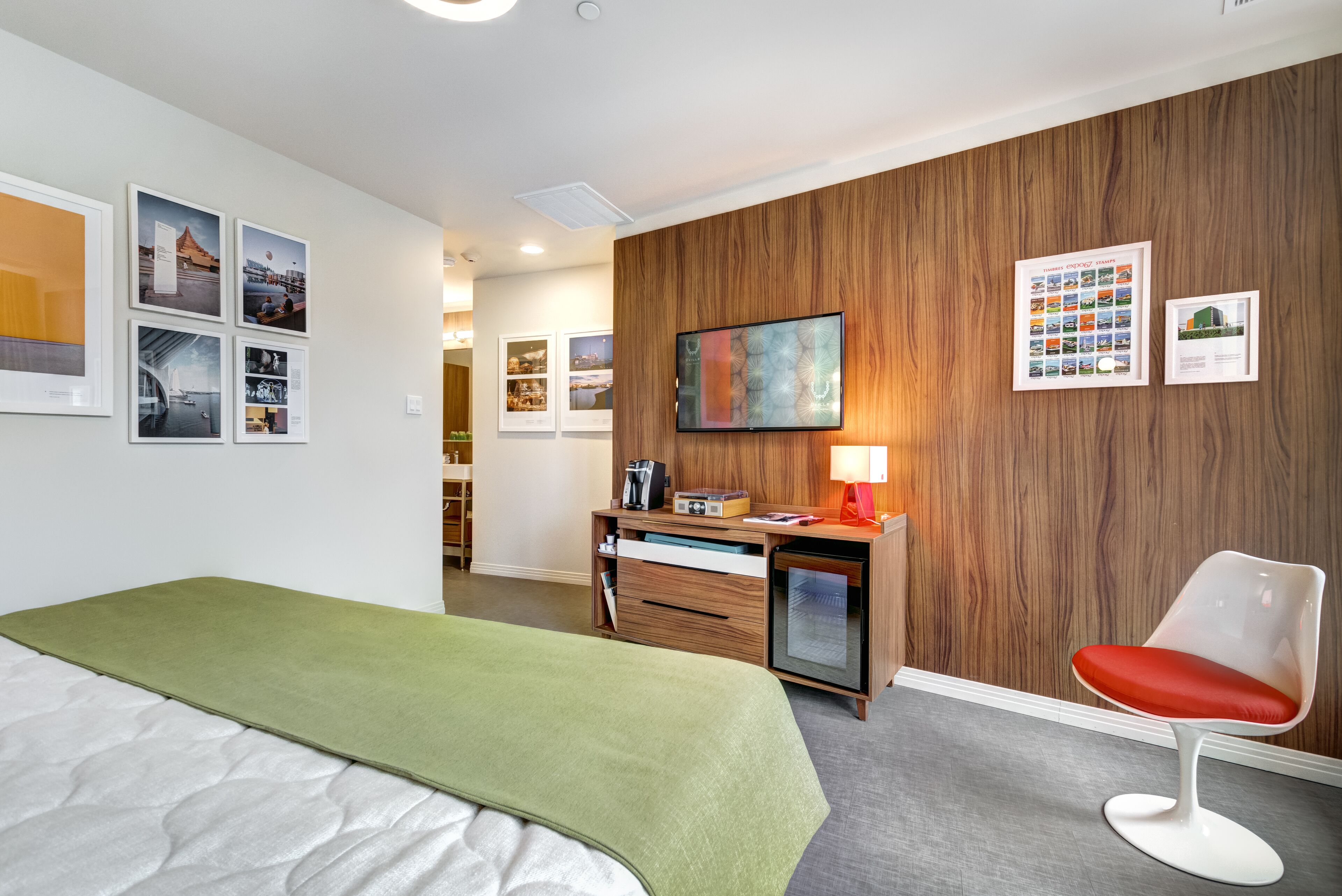 A hotel room with a bed, a desk with a lamp, a chair, and a wall with framed pictures and a television.