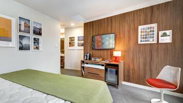 A hotel room with a bed, a desk with a lamp, a chair, and a wall with framed pictures and a television.