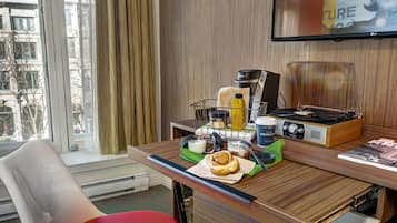 A hotel room with a wooden desk, a coffee maker, a radio, a bottle, and a pastry.