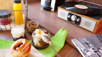 Uma mesa de madeira com toca-discos de vinil, um pote de mix de frutos secos, um copo de iogurte, um pastel e uma garrafa de sumo de laranja.