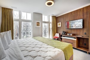 A hotel room with a large bed, a wooden desk, a television, and a view of the city through large windows.