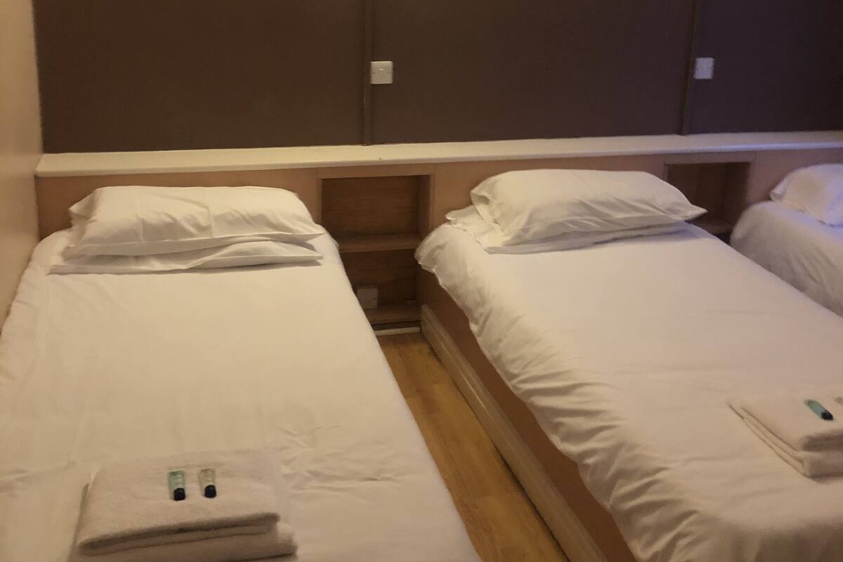 Habitación de hotel con dos camas individuales, cada una con almohada y manta. Hay una mesita de noche de madera entre las camas, y una lámpara de pared sobre cada cama.