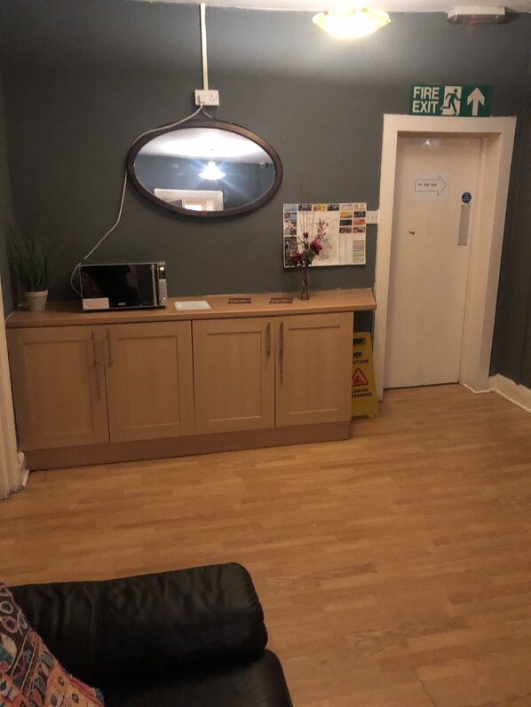 A room with a wooden cabinet, a microwave, a mirror, a fire exit sign, and a door with a caution sign. - The Salford Arms Hotel - Adults Only (Salford)