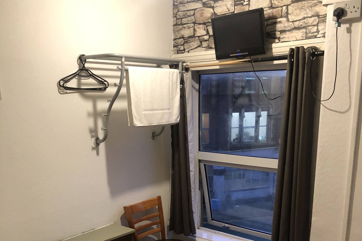 Una habitación de hotel con cama, una silla, una ventana con cortinas, un televisor de pantalla plana montado sobre la ventana y un perchero con una toalla.