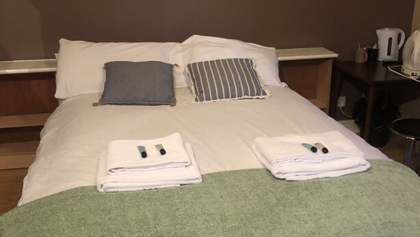 Una cama bien hecha con almohadas, un edredón verde y toallas dobladas. Se observa una mesita de noche con una tetera y una taza.