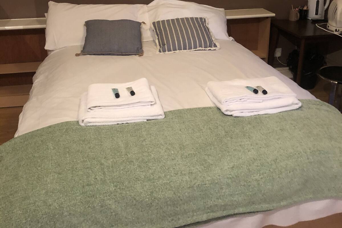 Una cama bien hecha con almohadas, un edredón verde y toallas dobladas. Se observa una mesita de noche con una tetera y una taza.