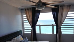 Een slaapkamer met een bed, een plafondventilator en uitzicht op de oceaan door een raam met gordijnen.