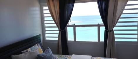 A bedroom with a bed, a ceiling fan, and a view of the ocean through a window with curtains.