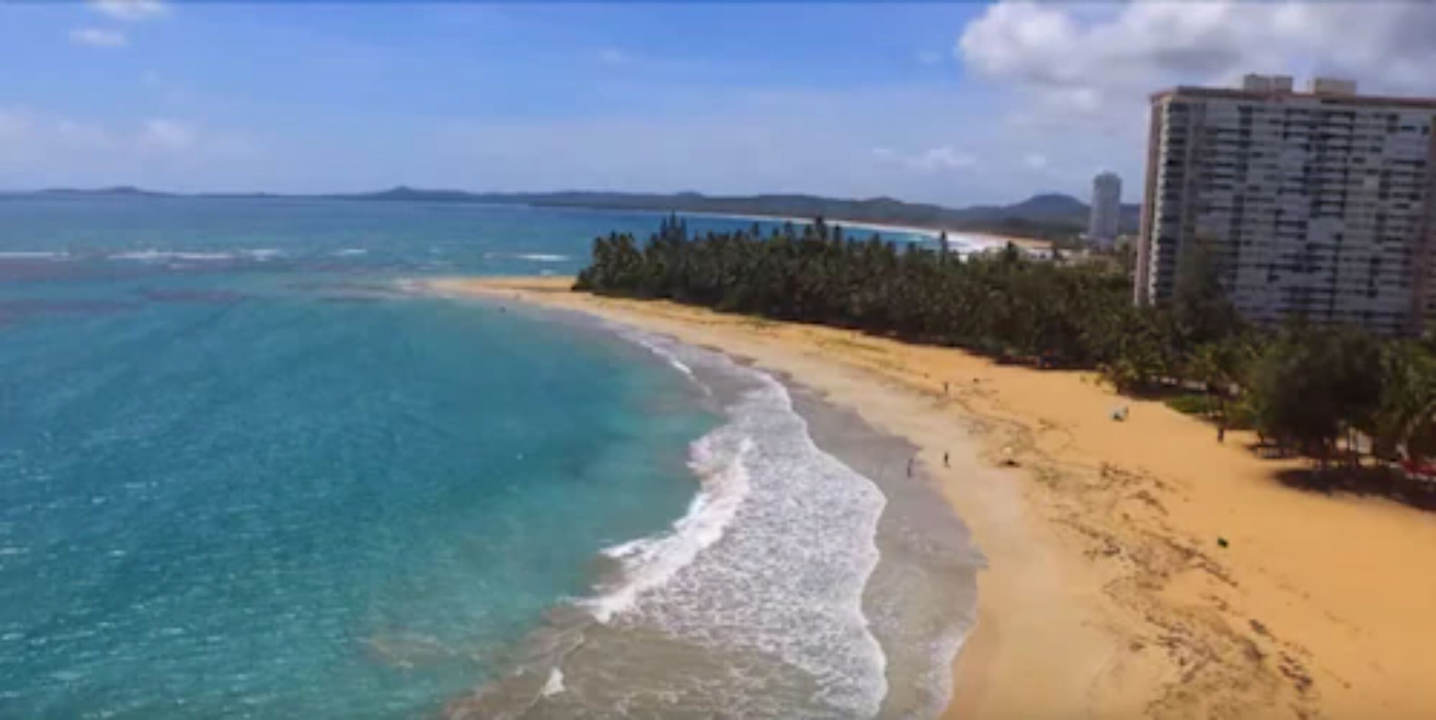 Una playa con agua turquesa cristalina, una orilla de arena y un edificio alto al fondo.