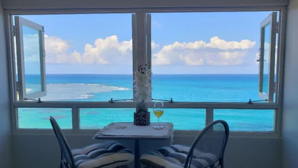 Een kamer met een tafel en twee stoelen, gericht op een groot raam met uitzicht op een strand en de oceaan.