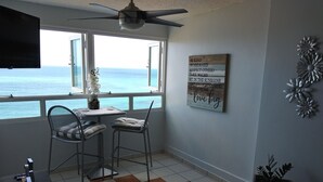 Een kamer met uitzicht op de oceaan, een plafondventilator, twee stoelen, een tafel met een vaas en een motiverende quote op de muur.