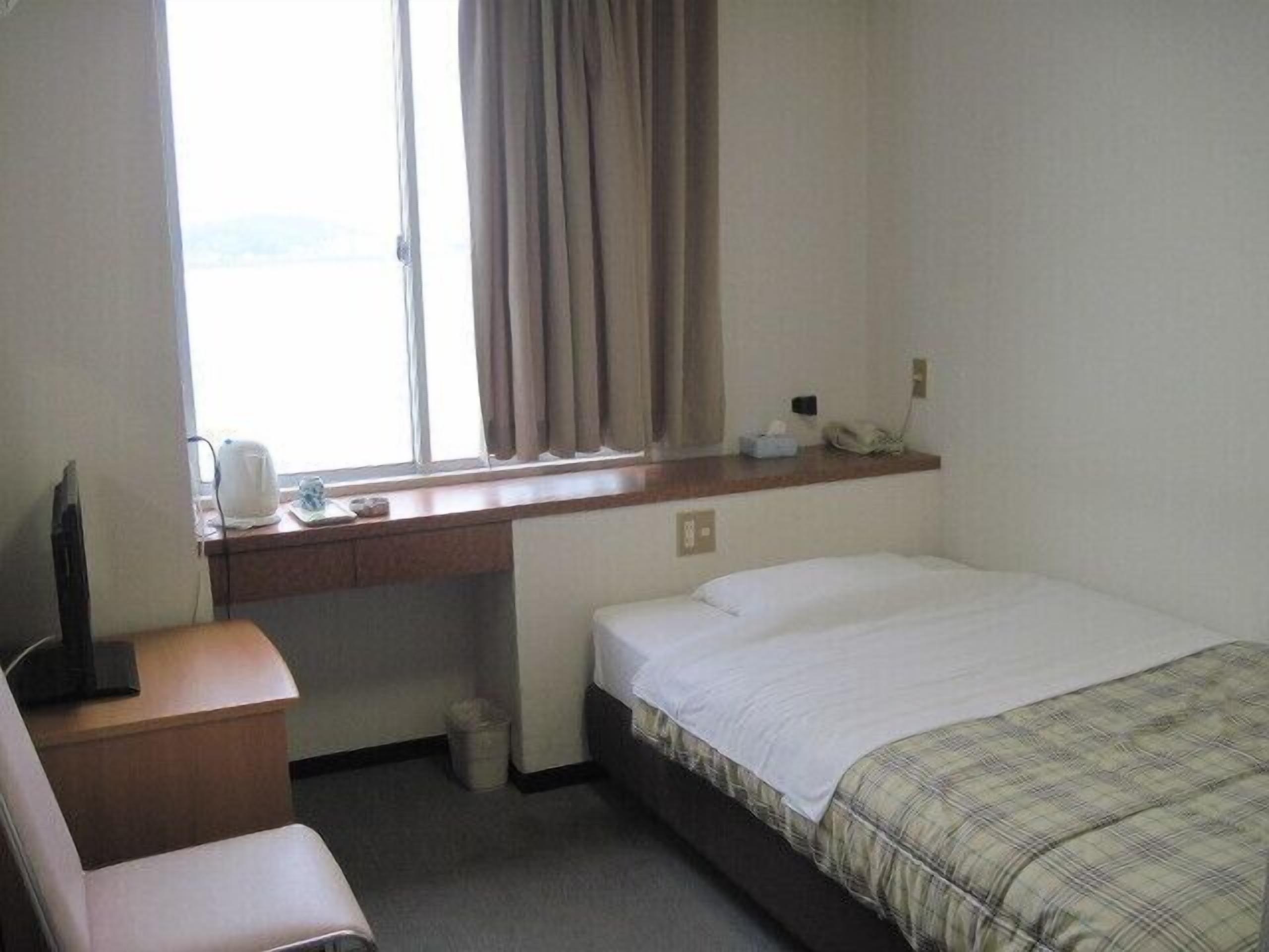 A hotel room with a bed, a desk with a telephone, a chair, a window with curtains, and a small trash bin.