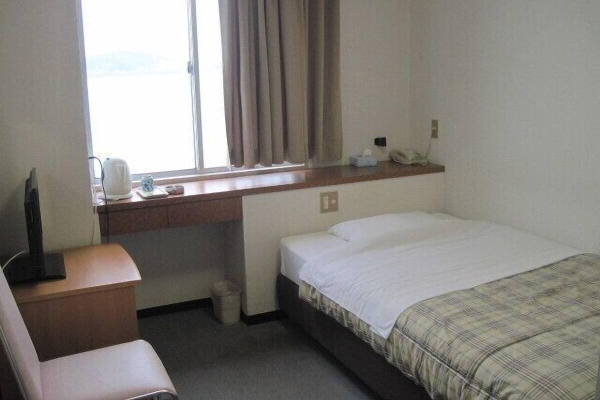 A hotel room with a bed, a desk with a telephone, a chair, a window with curtains, and a small trash bin.
