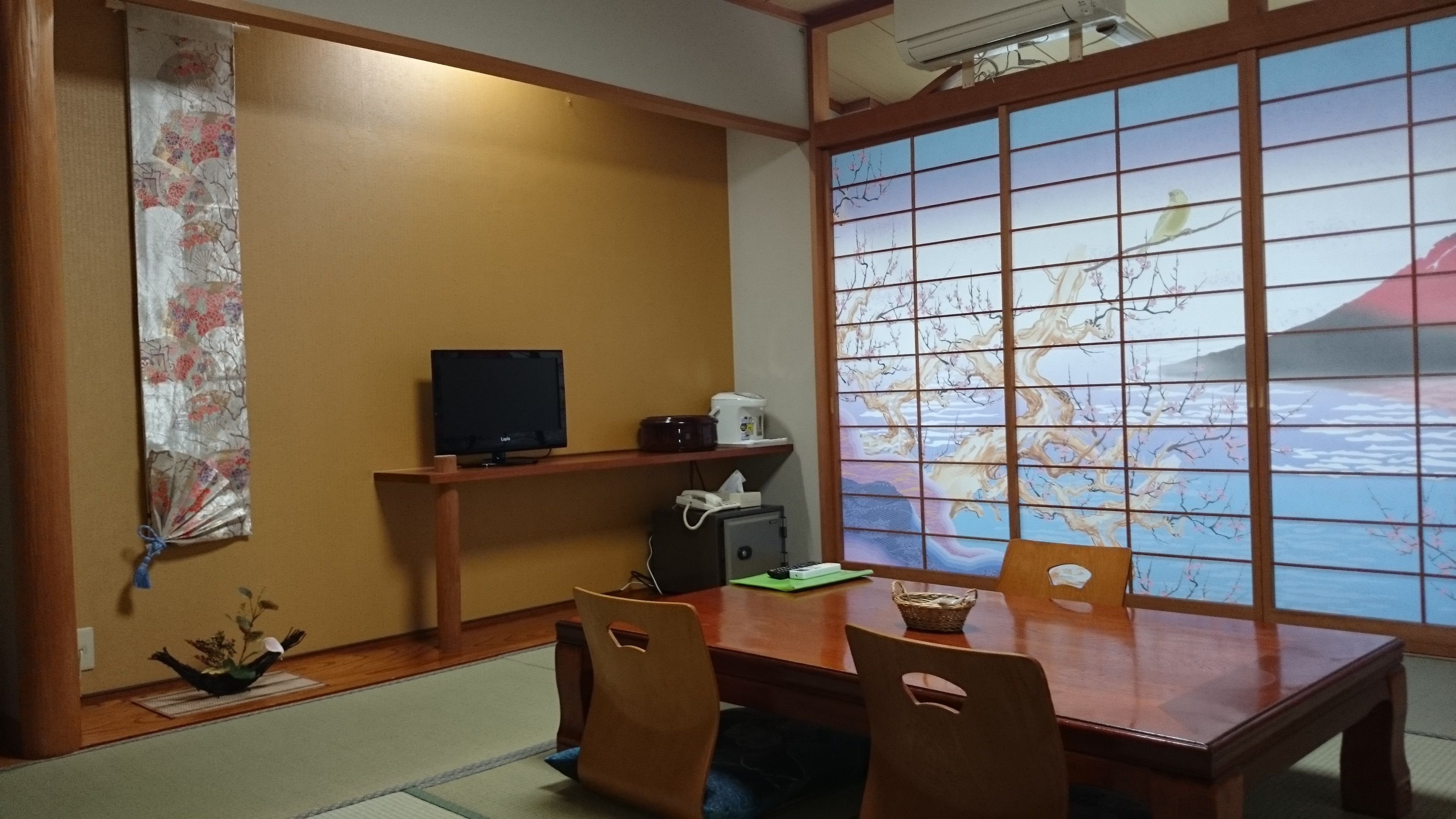 A traditional Japanese room with tatami flooring, a low table with chairs, a television, a small heater, and a view of cherry blossoms through a shoji screen.