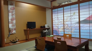 A traditional Japanese room with tatami flooring, a low table with chairs, a television, a small heater, and a view of cherry blossoms through a shoji screen.