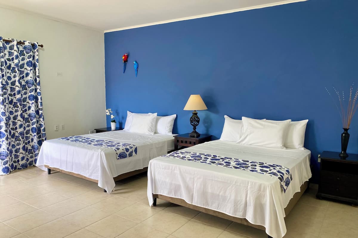 A hotel room with two beds, a blue accent wall, a nightstand with a lamp, and a decorative plant.