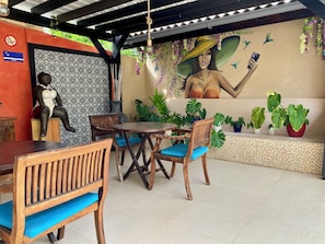 Un área de estar al aire libre con techo, un mural, muebles de madera y plantas en macetas.