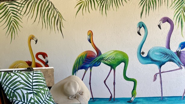 Une pièce décorée d’une fresque représentant des flamants roses aux couleurs vives, un canapé vert agrémenté d’un coussin à motifs et une table en bois dont le plateau est aux couleurs de l’arc-en-ciel.