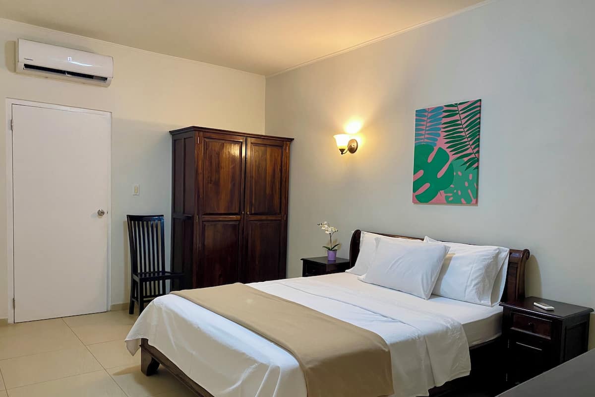 A hotel room with a bed, a wardrobe, a nightstand, a wall-mounted lamp, and a door.