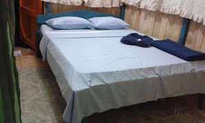 Una cama individual con cabecera azul y ropa de cama blanca en una habitación con paredes de madera y suelo de baldosas.