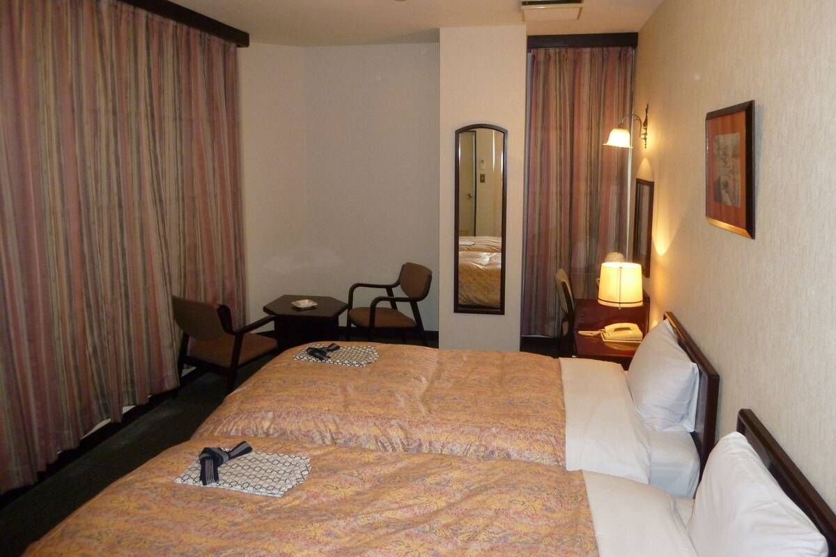 A hotel room with a double bed, two chairs, a small table, a lamp, a mirror, and a painting on the wall.