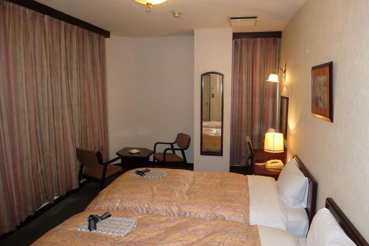 A hotel room with a double bed, a desk with a chair, a small table, a lamp, a mirror, and curtains.