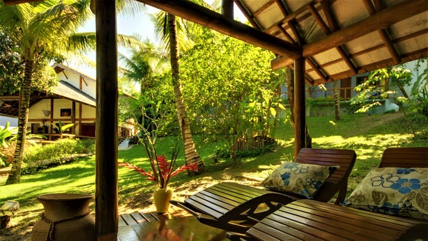 A wooden deck with a bench, a vase with red flowers, and a house in the background. - Atmosphera Pousada (Porto Seguro)