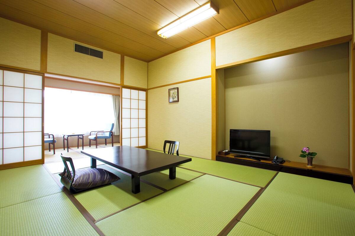 A traditional Japanese room with tatami flooring, a low table, chairs, a television, and a small plant.
