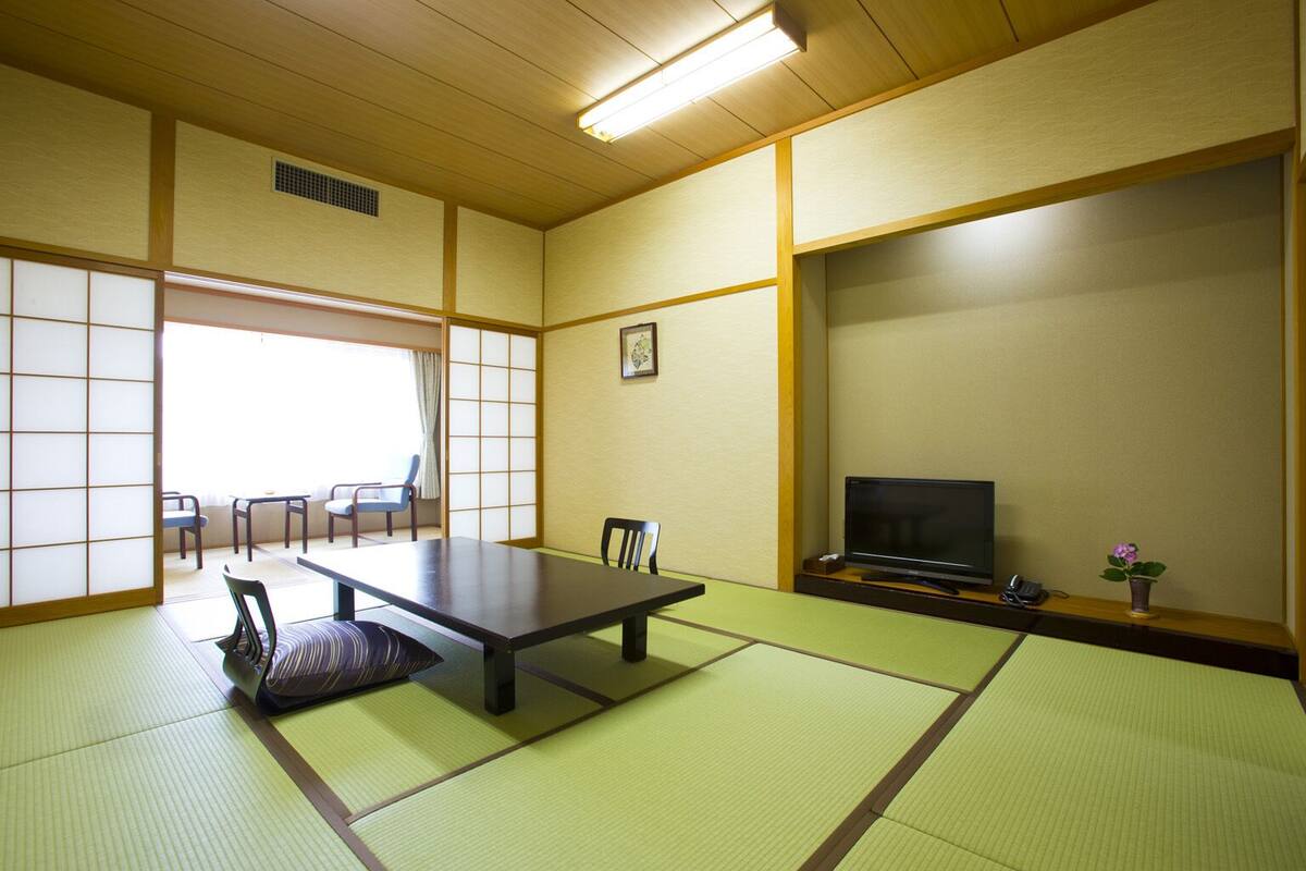 A traditional Japanese room with tatami flooring, a low table, chairs, a television, and a small plant.