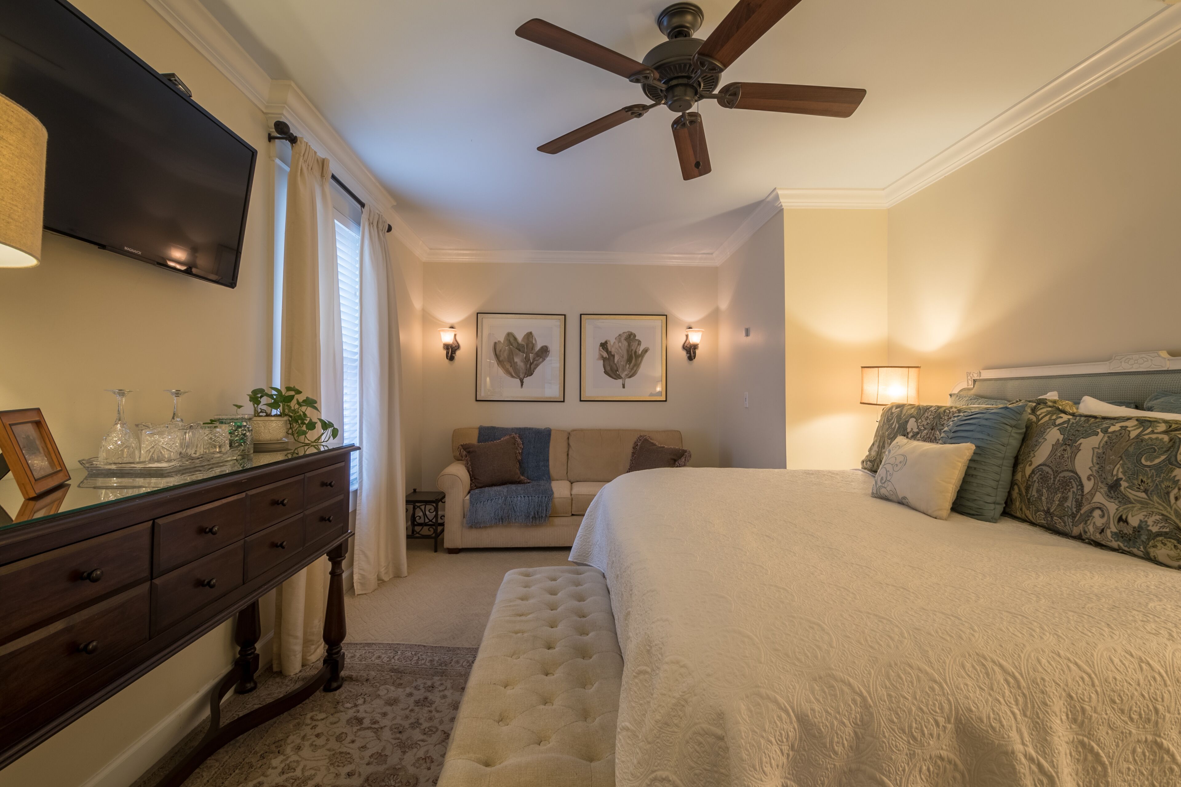 Une chambre à coucher comprenant un lit, un ventilateur de plafond, une commode, un téléviseur et des lampes fixées au mur.