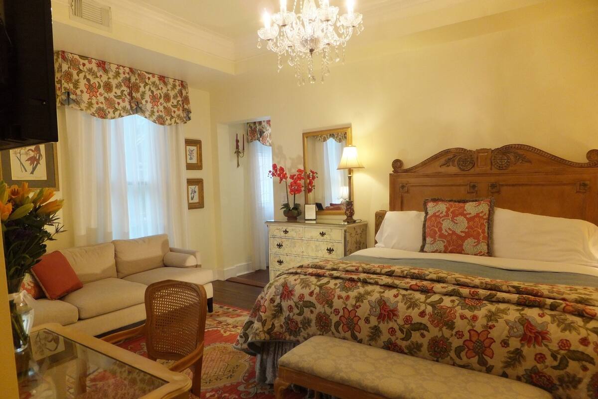 A hotel room with a bed, a sofa, a chair, a dresser, and a chandelier.