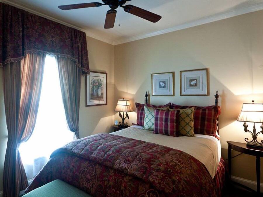 Una habitación de hotel con una cama, un ventilador de techo, dos lámparas de noche y un ventanal con cortinas.