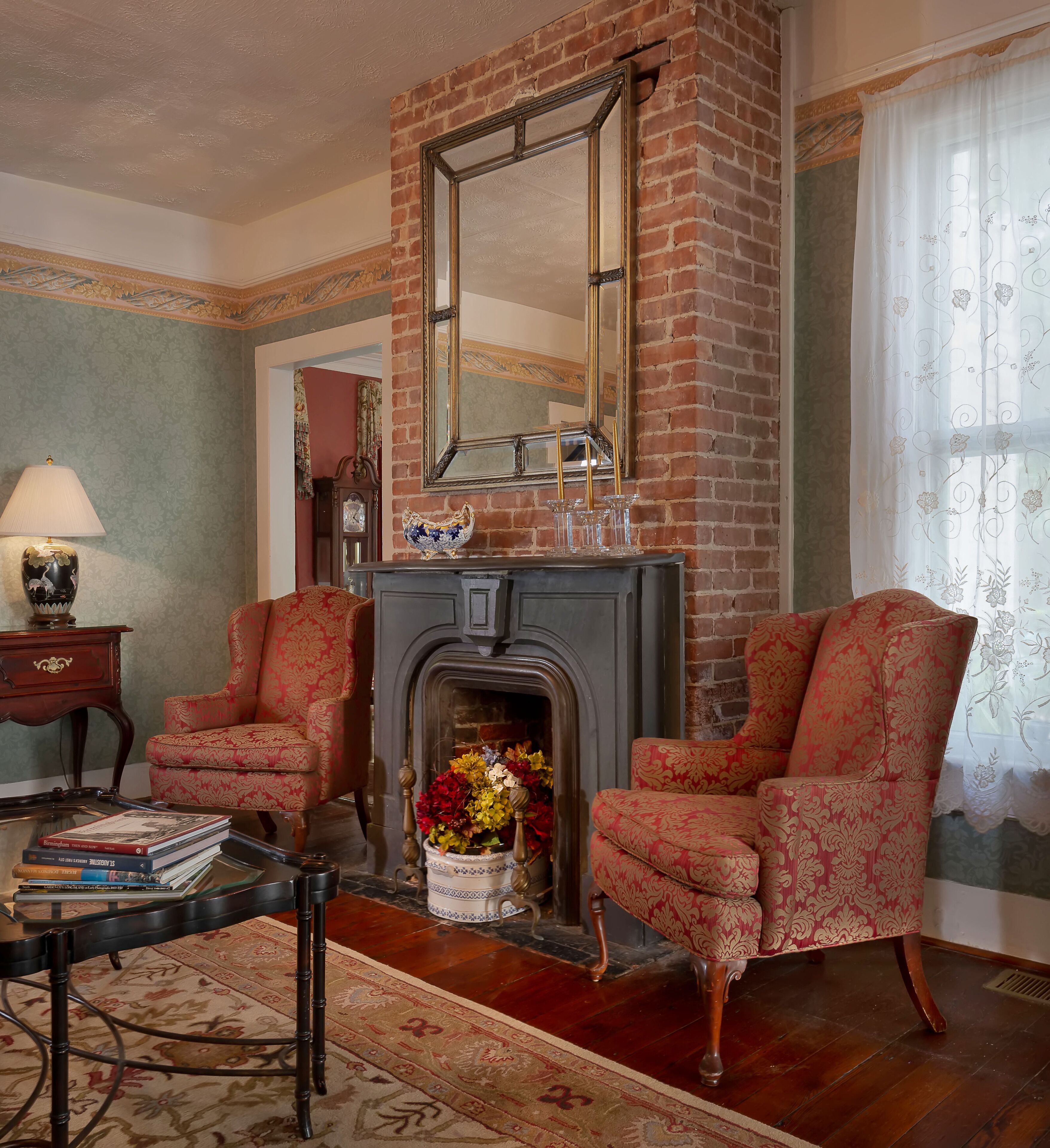 A traditional living room with a brick fireplace, two armchairs, a coffee table, and a bookshelf.