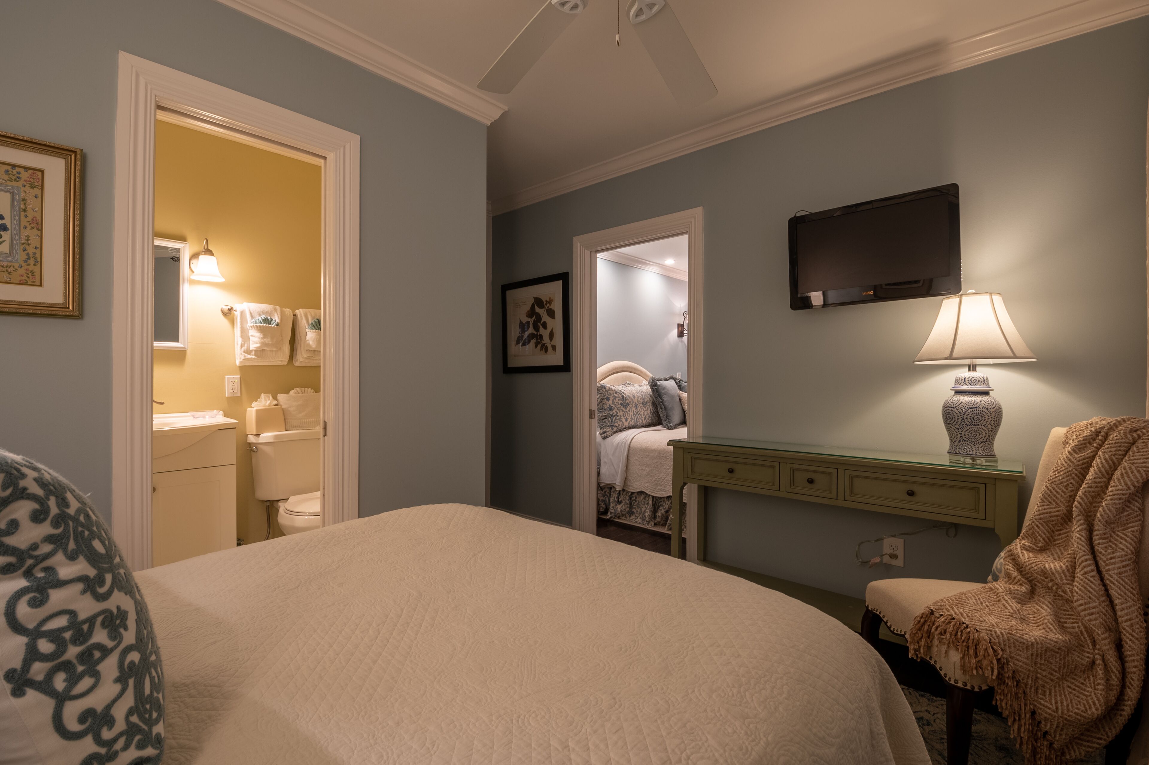 A bedroom with a large bed, a bedside table with a lamp, a chair with a blanket, a TV mounted on the wall, and a bathroom visible through an open door.
