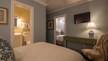 A bedroom with a large bed, a bedside table with a lamp, a chair with a blanket, a TV mounted on the wall, and a bathroom visible through an open door.