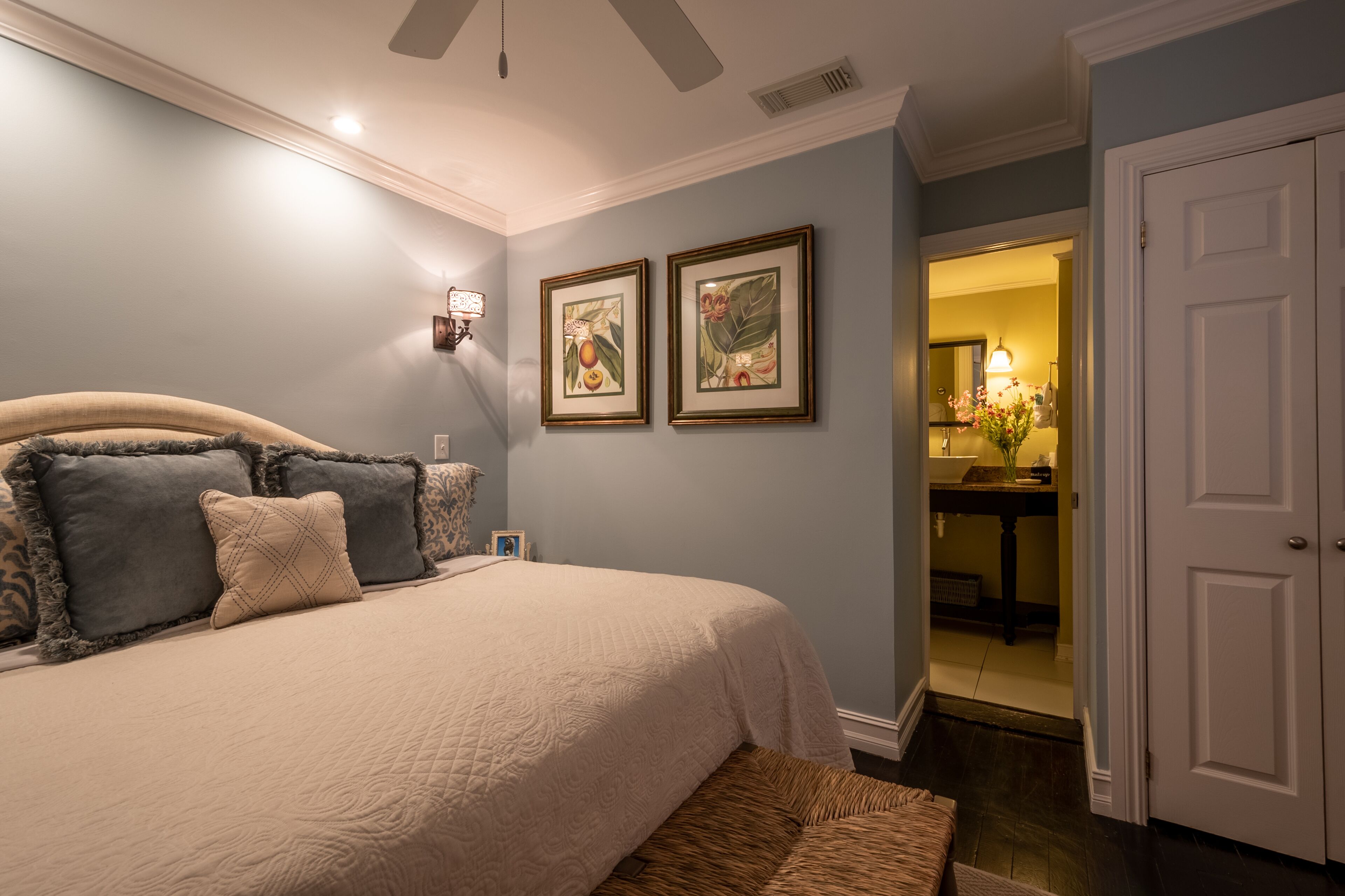 A bedroom with a bed, pillows, a nightstand, a wall-mounted lamp, and two framed pictures.