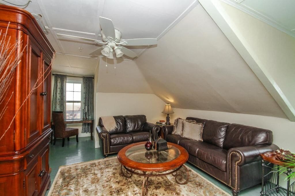 Een ruime woonkamer met een hoge plafond, een plafondventilator, een groot raam, een bruine leren bank, een houten salontafel en een donker houten kast.