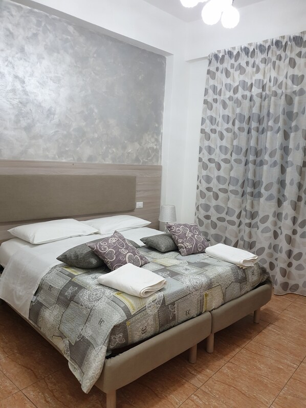 A neatly made bed with patterned bedding and pillows, a bedside lamp, and a large window with patterned curtains.