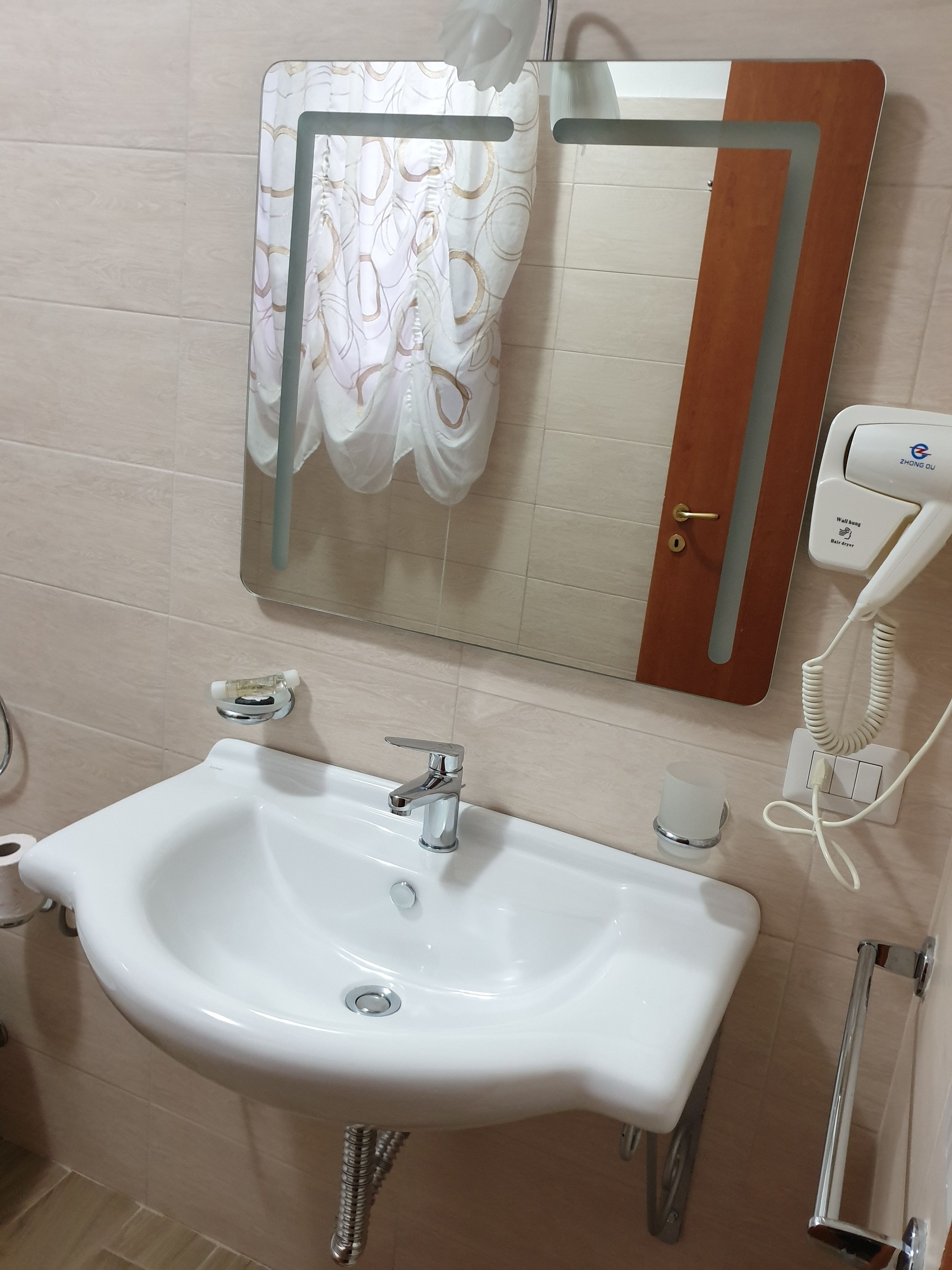 A bathroom with a white sink, a mirror, a towel rack with a white towel, a toilet paper holder, and a hand dryer.