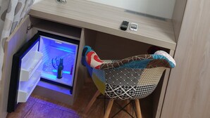 A compact room with a TV mounted on the wall, a small shelf with a vase and remote controls, a chair with a colorful cushion, and a mini fridge with a blue light. - Beauty house (Reggio Calabria)