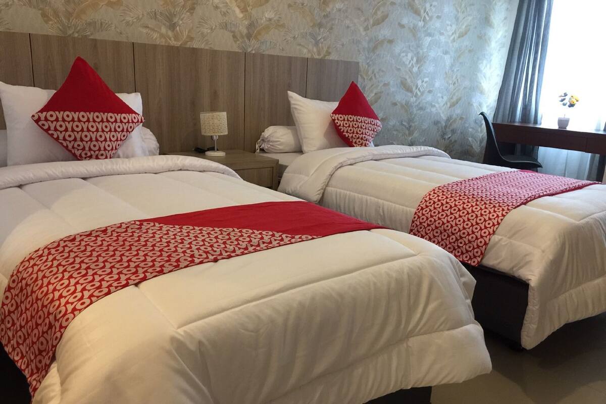 Habitación de hotel con dos camas, cada una con ropa blanca y detalles rojos. Tiene un cabecero de madera, una mesita de noche con lámpara y una silla.