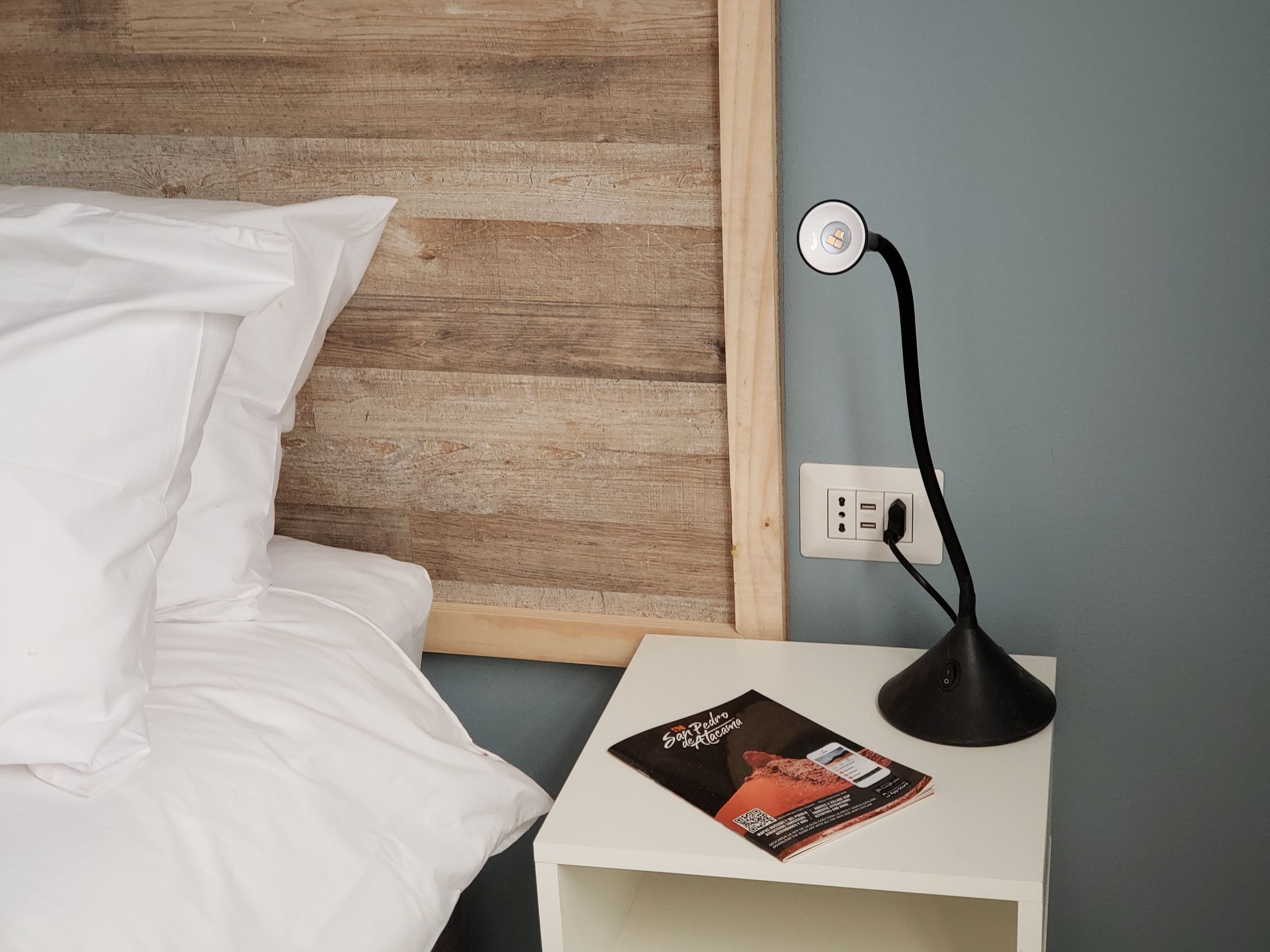 Una cama bien hecha con sábanas blancas, una mesita de noche con una lámpara y un folleto sobre la mesa.
