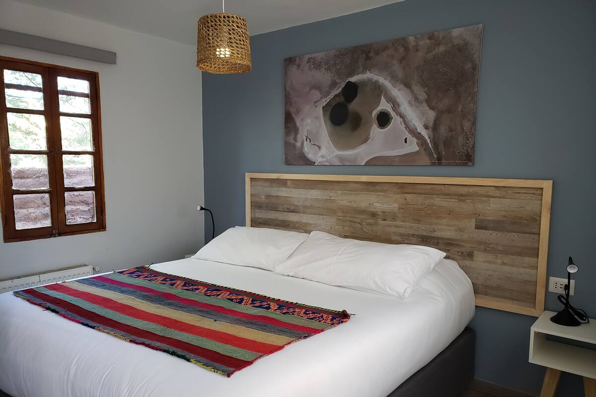 Een slaapkamer met een houten hoofdbord, wit beddengoed, een kleurrijke strepenworp, een aan de muur hangend abstract schilderij, een raam met uitzicht en een nachtkastje met een lamp.