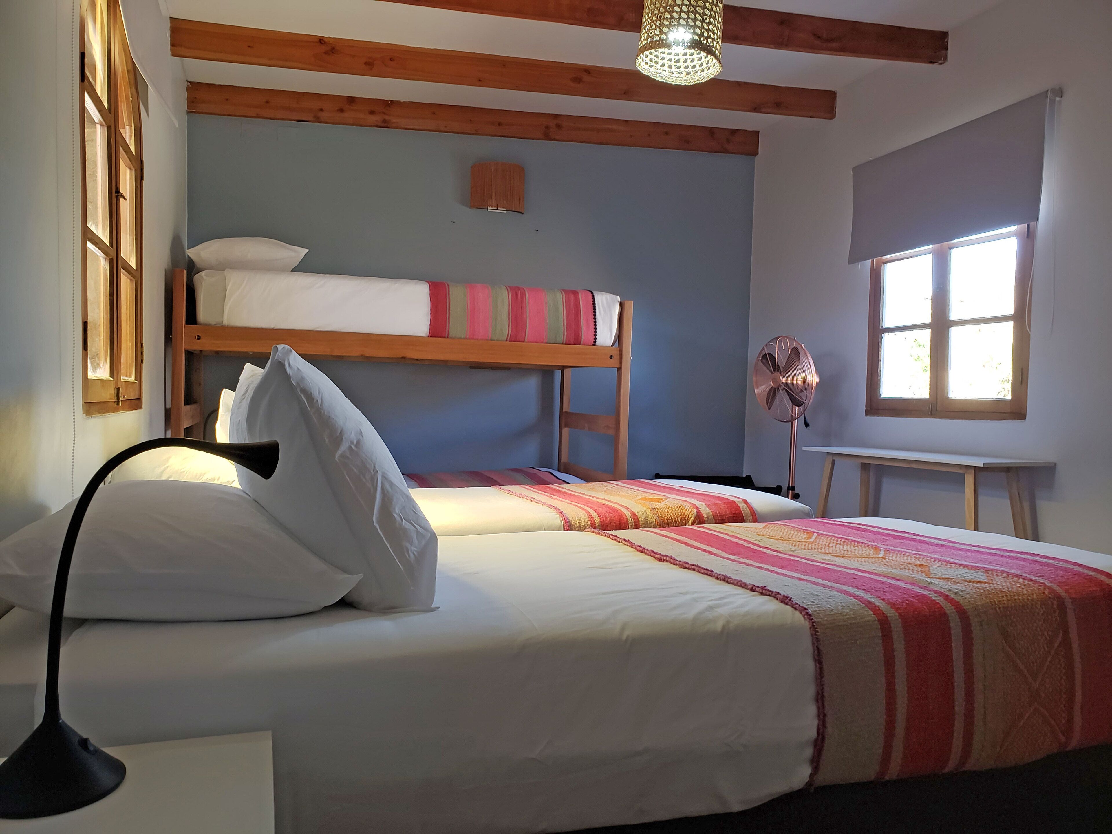 Un dormitorio con dos camas, un ventilador de pie, una ventana y vigas de madera en el techo.