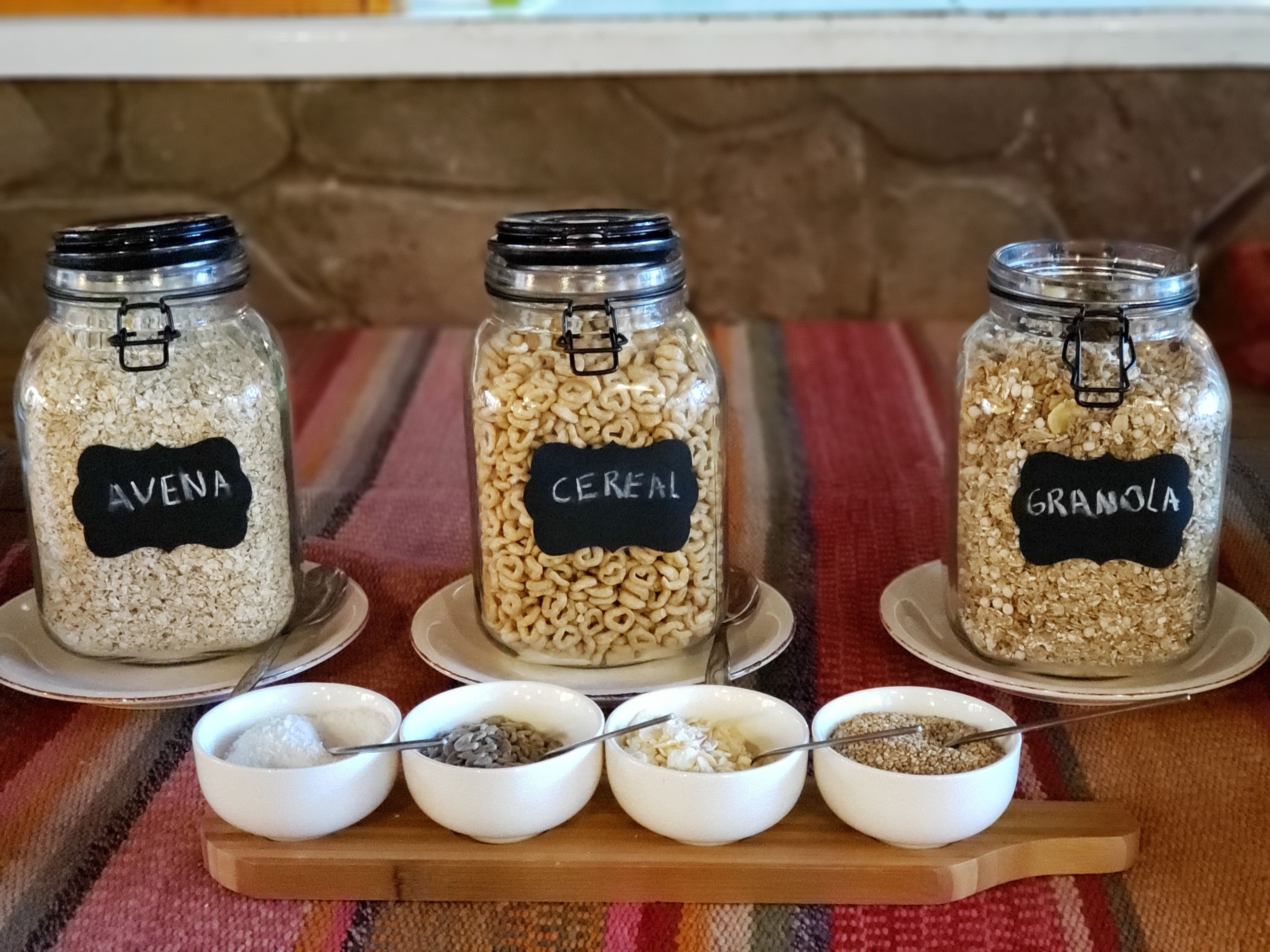 Three glass jars labeled 'AVENA,' 'CEREAL,' and 'GRANOLA' on a wooden board with bowls of grains in front.