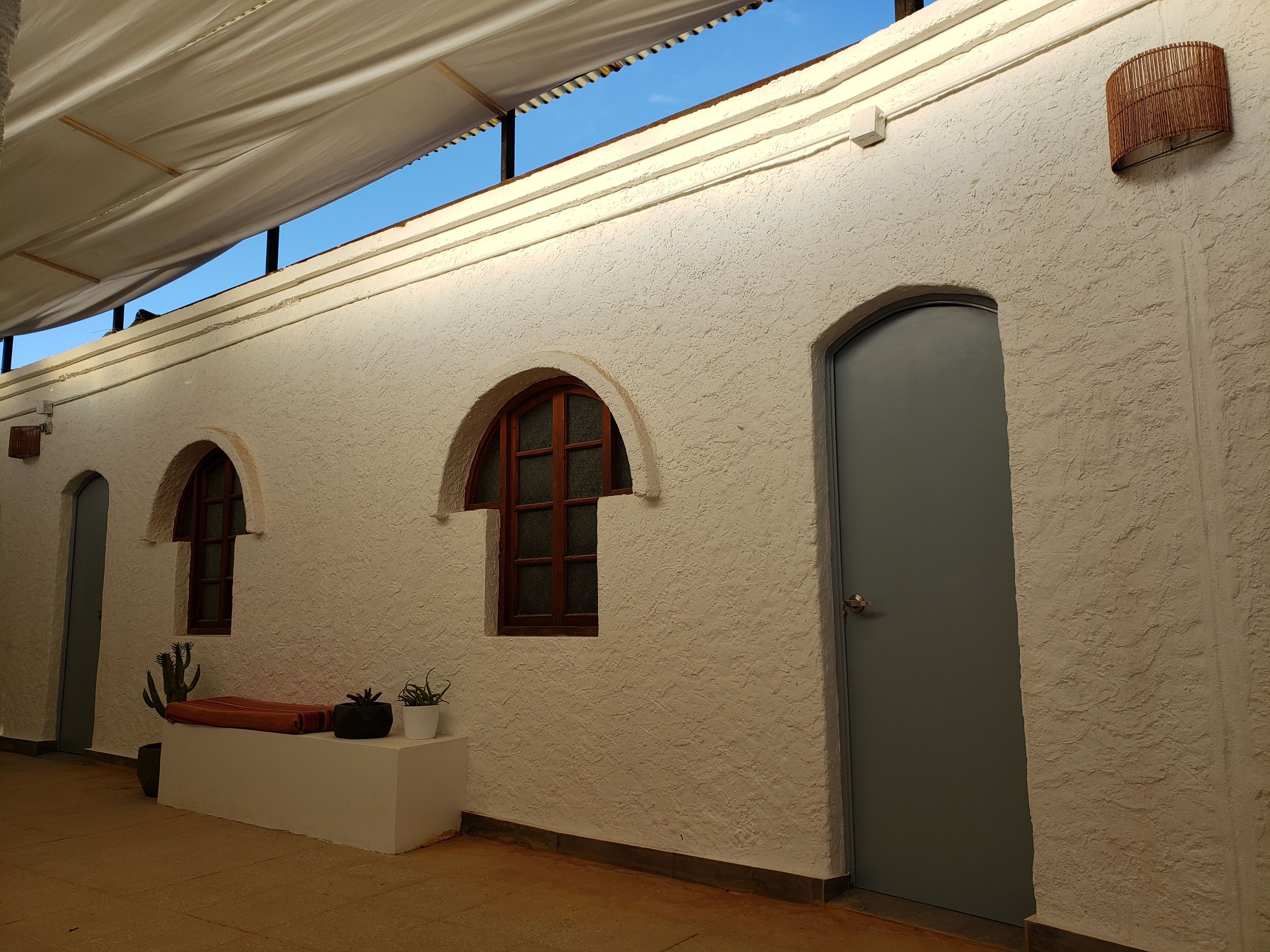 A corridor with white textured walls, three arched windows, a door, and a white bench with potted plants.